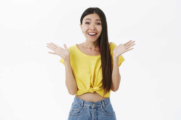 Erfreutes süßes asiatisches Mädchen mit langen dunklen Haaren im kurzen gelben T-Shirt, das Hände fasst und lächelt