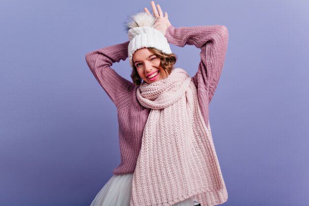 Erfreutes weibliches Modell mit glücklichem Gesichtsausdruck, der in Winterkleidung aufwirft und lächelt. Kurzhaarige Frau im Schal, die positive Gefühle auf lila Wand ausdrückt.