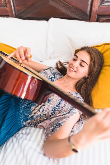 Kostenloses Foto erhöhte ansicht der glücklichen jugendlichen, die auf dem bett spielt gitarre liegt