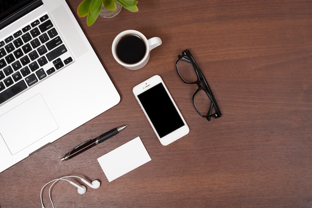 Erhöhte Ansicht des Laptops; Mobiltelefon; Tee; Ohrhörer; Brille mit Stift und Auge auf Holztisch