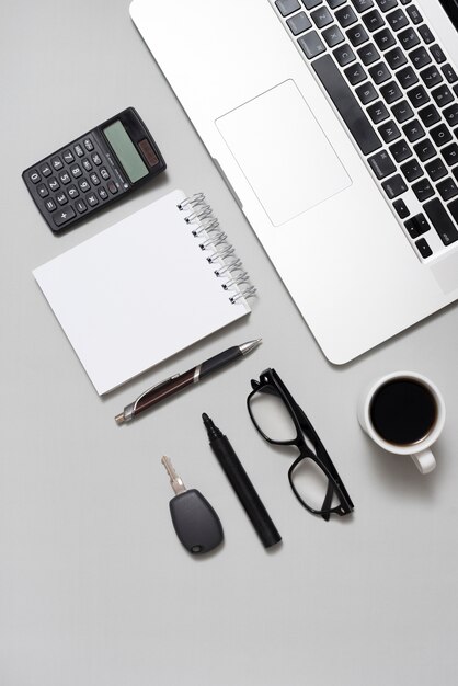 Erhöhte Ansicht des Laptops; Taschenrechner; leerer Notizblock; Brille; und Kaffeetasse mit Schlüssel auf grauem Hintergrund