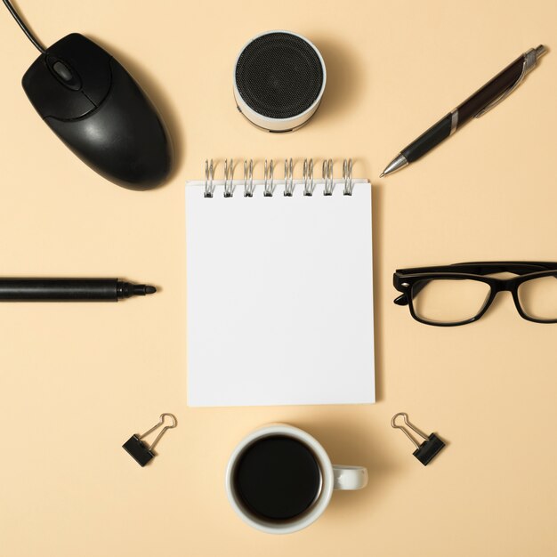 Erhöhte Ansicht des leeren Notizblatts umgeben von Bluetooth-Lautsprecher; Stift; Büroklammern; Kaffeetasse; Brille auf beige Hintergrund