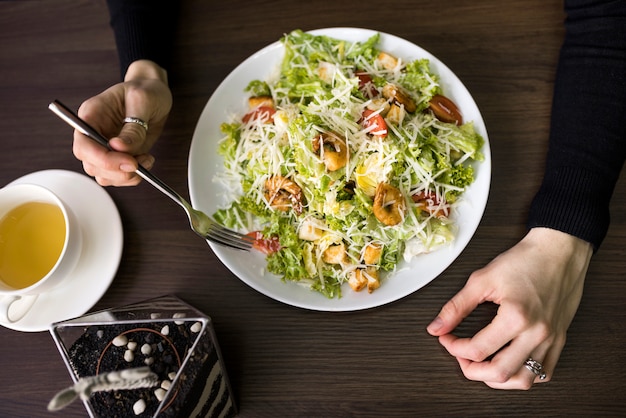 Erhöhte Ansicht einer Person, die Caesar-Salat mit Garnele auf weißer Platte über Tabelle isst