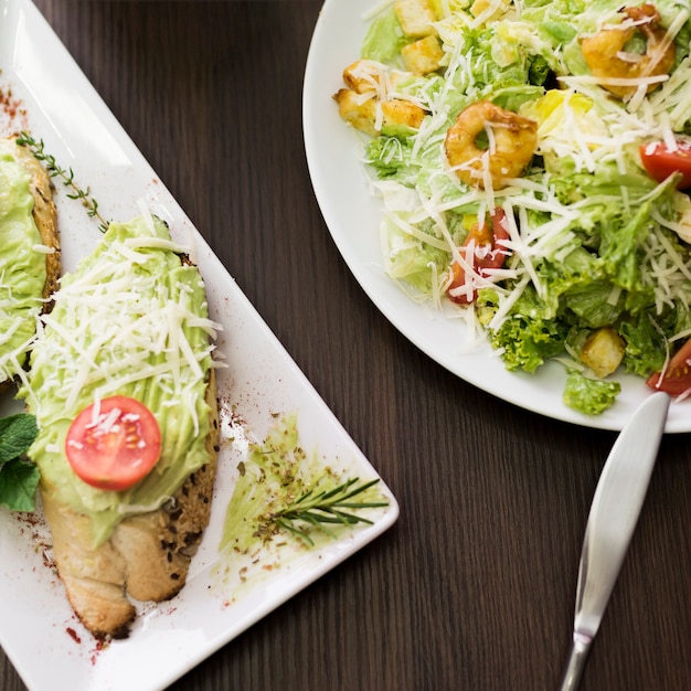 Erhöhte Ansicht von Brot mit Pesto-Sauce; geriebener Käse und Kirschtomate auf Teller in der Nähe von Salat über Tisch