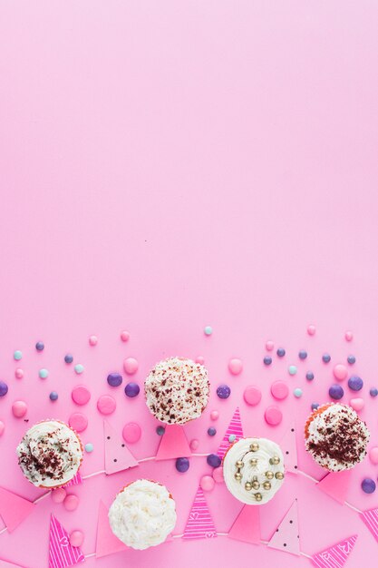 Erhöhte Ansicht von Cupcakes; Süßigkeiten und Fahne auf rosa Oberfläche