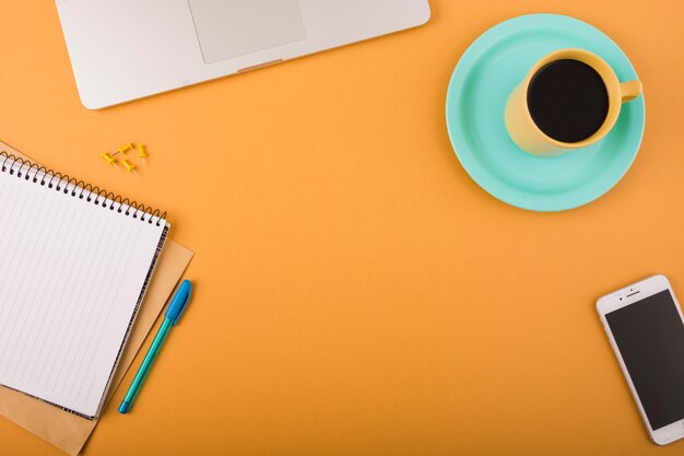 Erhöhte Ansicht von schwarzem Kaffee; Smartphone; Stift; Drucknadeln; Laptop und Notizblock auf orange Hintergrund