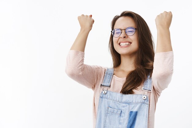 Erleichterte und glücklich aufgeregte Frau mit Brille, die breit lächelt, die geballten Fäuste zum Feiern und Triumph hebt, als Ziel jubelt und erfreut über grauer Wand posiert.