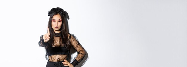 Ernst aussehende sexy Asiatin im schwarzen gotischen Kleid, die Halloween feiert und Stoppgeste sha zeigt