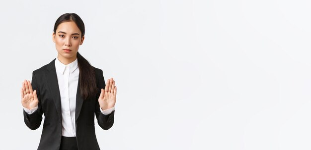 Ernst aussehende, unzufriedene asiatische Managerin, Geschäftsfrau im schwarzen Anzug, stoppt die Aktion, verbietet oder beschränkt etwas, das die Hände in einer verbotenen Geste hebt, die nein oder genügend weißen Hintergrund sagt
