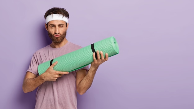 Kostenloses Foto ernst motivierter mann mit dicken borsten, hält aufgerollte kareamt, macht grimasse, bereit für yoga-training, trägt freizeitkleidung