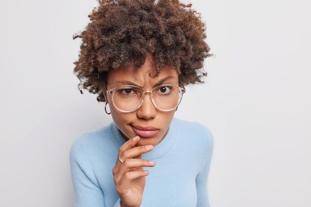 Kostenloses Foto ernste, aufmerksame frau mit lockigem haar hält kinntaschen, lippen, hat unzufriedenen ausdruck, trägt große brillen, lässiger blauer pullover, isoliert auf weißem hintergrund, hält etwas für streng