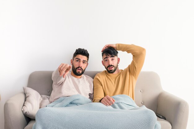 Ernste homosexuelle Paare, die auf Sofa gegen die weiße Wand fernsehen sitzen