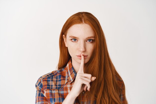 Ernste junge Dame mit rotem Naturhaar, vorn verstummend, mit selbstbewusstem Gesicht ein Geheimnis verratend, über weißer Wand stehend