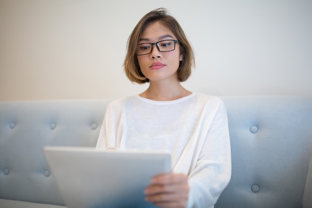 Ernste junge Frau, die auf Tablet-Computer auf Sofa grast