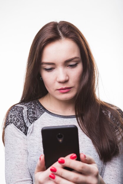 Ernste junge Frau, die ihren Smartphone auf weißem Hintergrund betrachtet