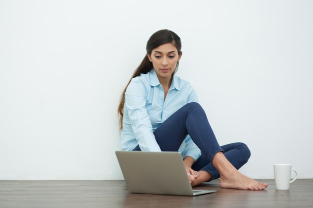 Ernste junge Frau mit Laptop und Tee auf Boden