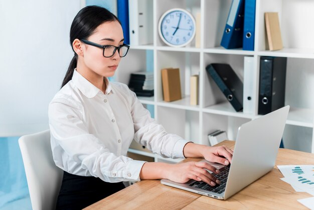 Ernste junge Geschäftsfrau, die auf Laptop über dem Schreibtisch schreibt