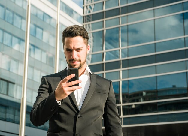 Ernste junge Geschäftsfrau, die den Handy steht in der Front des Unternehmensgebäudes betrachtet