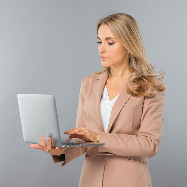 Ernste junge Geschäftsfrau, die Laptop auf ihrer Hand gegen grauen Hintergrund verwendet