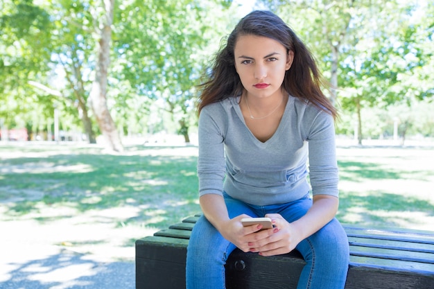 Ernste recht junge Dame, die Smartphone auf Bank im Park verwendet