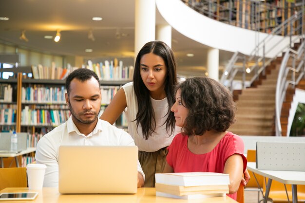 Ernste Studenten, die bei Tisch in der Bibliothek arbeitet mit Laptop sitzen