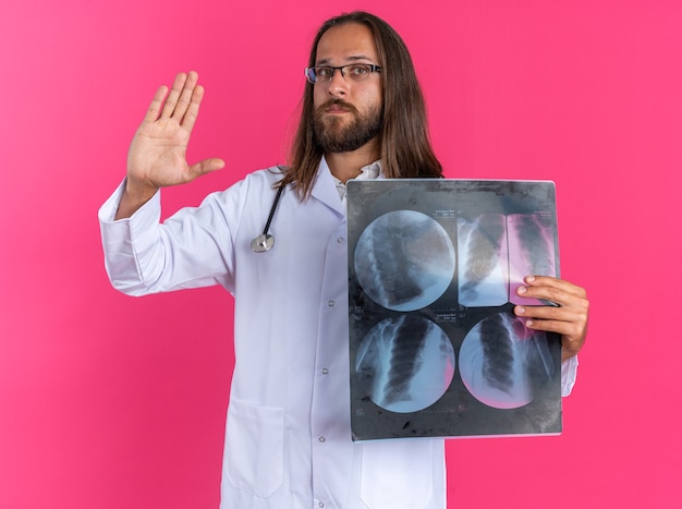 Ernster erwachsener männlicher Arzt, der medizinische Robe und Stethoskop mit Brille trägt, die eine Röntgenaufnahme zeigt, die auf die Kamera schaut und die Stoppgeste isoliert auf rosa Wand macht