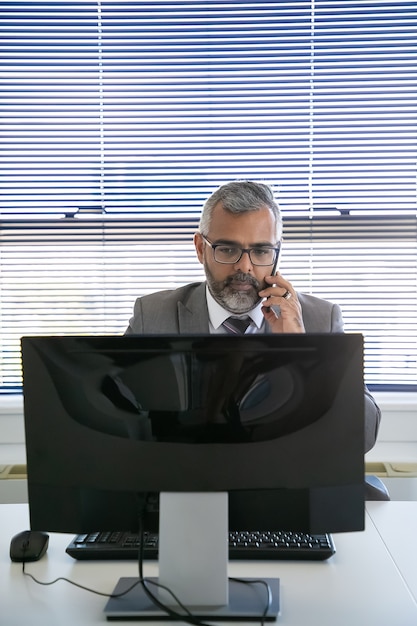 Ernster grauhaariger Geschäftsmann, der Anruf auf Handy macht, während Computer am Arbeitsplatz im Büro verwendet wird. Vorderansicht. Kommunikations- und Multitasking-Konzept