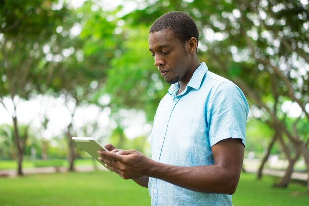 Ernster hübscher junger afrikanischer Kerl, der Tablette im Sommerpark verwendet.