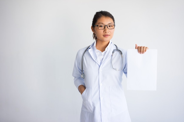 Ernster junger asiatischer weiblicher Doktor, der leeres Blatt Papier zeigt.