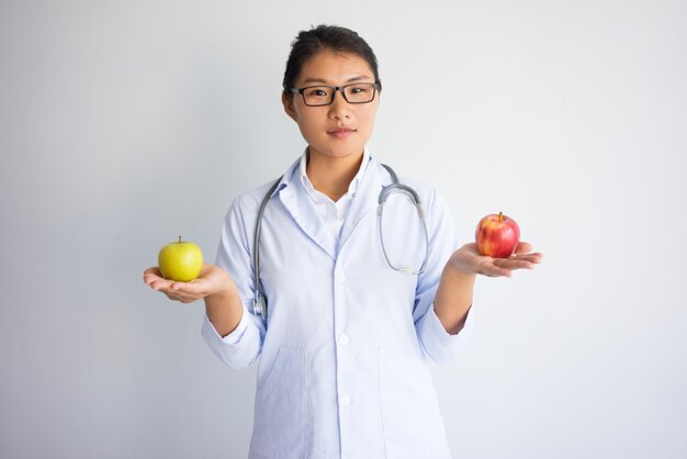 Ernster junger asiatischer weiblicher Doktor, der roten und gelben Apfel anhält.