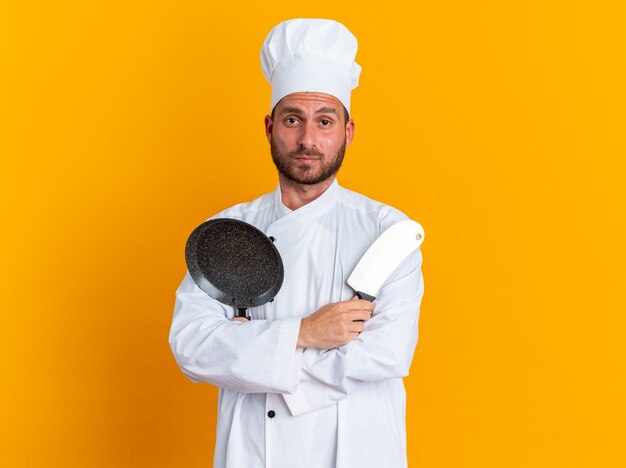 Ernster junger kaukasischer männlicher Koch in Kochuniform und Mütze stehend mit geschlossener Haltung mit Hackmesser und Bratpfanne mit Blick auf die Kamera isoliert auf oranger Wand mit Kopierraum