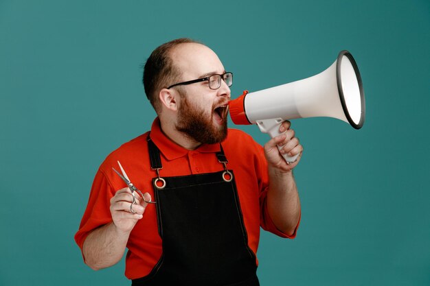 Ernster junger männlicher Friseur mit Brille, rotem Hemd und Friseurschürze mit einer Schere, die seitlich in den Lautsprecher spricht, isoliert auf blauem Hintergrund