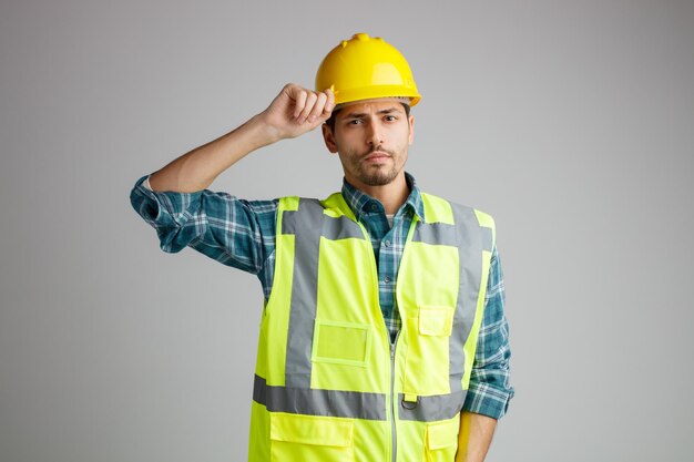 Ernster junger männlicher Ingenieur mit Schutzhelm und Uniform, der in die Kamera blickt, während er seinen Helm isoliert auf weißem Hintergrund ergreift