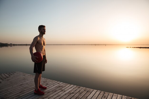 Ernster junger Sportler, der mit Ball am Strand steht.