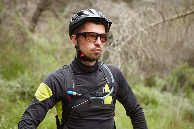 Ernster kaukasischer männlicher Radfahrer, der Schutzhelm und -brille trägt, die sich nach intensivem Radsporttraining im Freien im Wald auf seinem Booster-Fahrrad müde fühlen