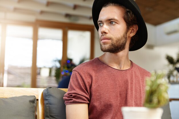 Ernster oder trauriger attraktiver junger bärtiger Student, der trendigen schwarzen Hut trägt, der allein im modernen geräumigen Café sitzt