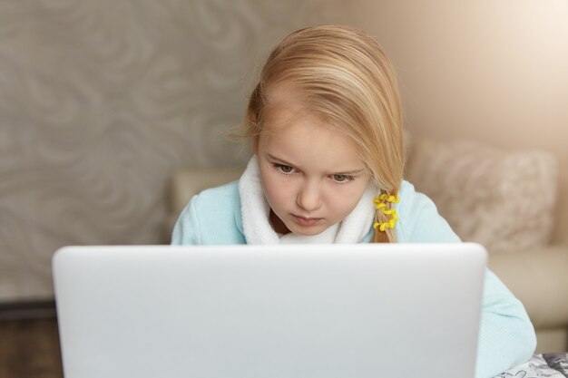 Ernstes blondes kleines Mädchen, das vor offenem Laptop sitzt