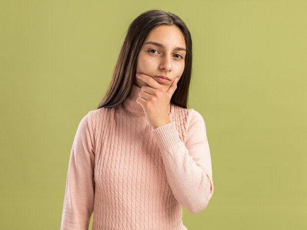 Ernstes hübsches Teenager-Mädchen, das die Kamera anschaut und die Hand am Kinn hält, isoliert auf olivgrüner Wand mit Kopienraum?