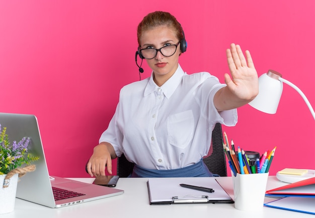 Ernstes junges blondes Call-Center-Mädchen mit Headset und Brille, das am Schreibtisch mit Arbeitswerkzeugen sitzt und Stopp-Geste macht