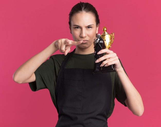 Ernstes junges brünettes Friseurmädchen in Uniform, das Haarschneidemaschinen und Siegerpokal hält und auf die Kamera zeigt