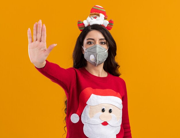 Ernstes junges hübsches Mädchen mit Weihnachtsmann-Stirnband und Pullover mit Schutzmaske, die Stopp-Geste einzeln auf orangefarbener Wand mit Kopierraum macht