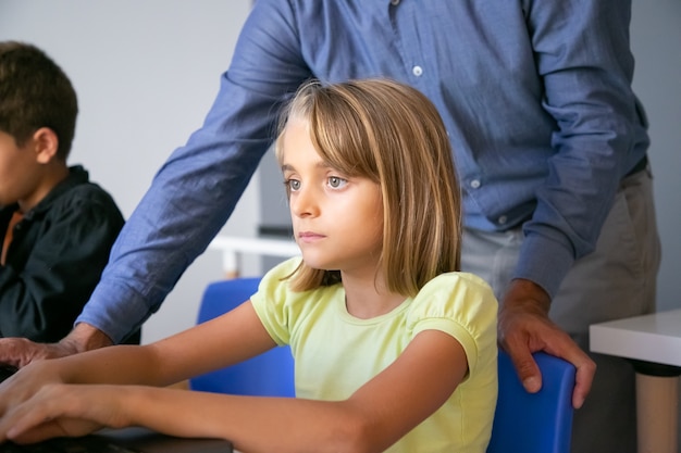 Ernstes kaukasisches Mädchen, das am Tisch im Klassenzimmer sitzt, Text auf dem Bildschirm liest oder Videopräsentation sieht