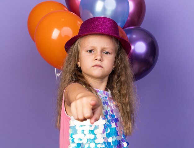 ernstes kleines blondes Mädchen mit violettem Partyhut, der vor Heliumballons zeigt, die auf lila Wand mit Kopienraum lokalisiert werden