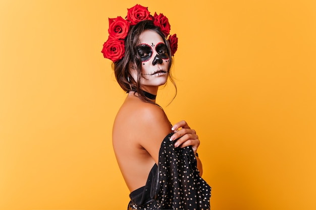 Ernstes Mädchen in la muerta Kleidung, die Kamera während Halloween-Fotoshooting schaut. Charmante tote Braut mit Rosen im schwarzen Haar lokalisiert auf gelbem Hintergrund.