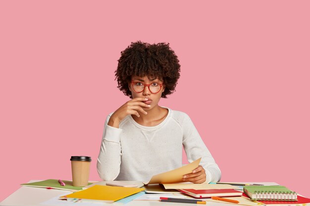 Ernstes Studentenmädchen, das am Schreibtisch gegen die rosa Wand aufwirft