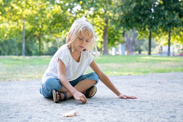 Ernstes süßes blondes Mädchen, das mit bunten Kreidestücken sitzt und zeichnet. Speicherplatz kopieren. Konzept für Kindheit und Kreativität