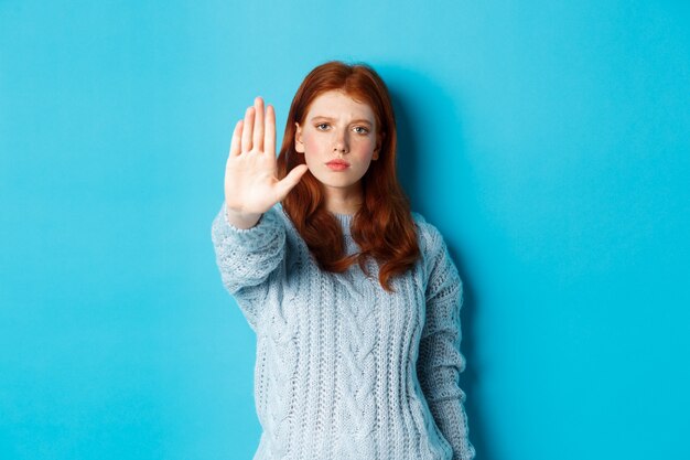 Ernstes und selbstbewusstes rothaariges Mädchen, das sagt, dass es aufhören soll, nein sagt, ausgestreckte Handfläche zeigt, um Aktionen zu verbieten, auf blauem Hintergrund stehend