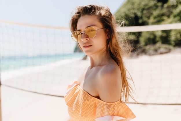 Ernstes weißes weibliches Modell in der Sonnenbrille, die am Strand aufwirft und schaut. Foto im Freien des nachdenklichen brünetten Mädchens in der orange Kleidung, die vor Volleyball-Set steht.