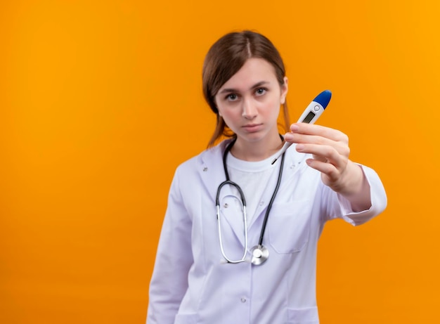 Ernsthaft aussehende junge Ärztin, die medizinische Robe und Stethoskop trägt und Thermometer auf lokalisiertem orange Raum mit Kopienraum streckt
