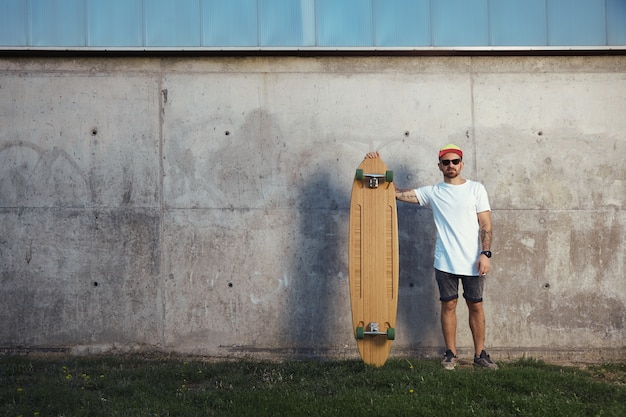 Ernsthaft aussehender Surfer mit Bart, Tätowierungen und Sonnenbrille neben seinem Longboard